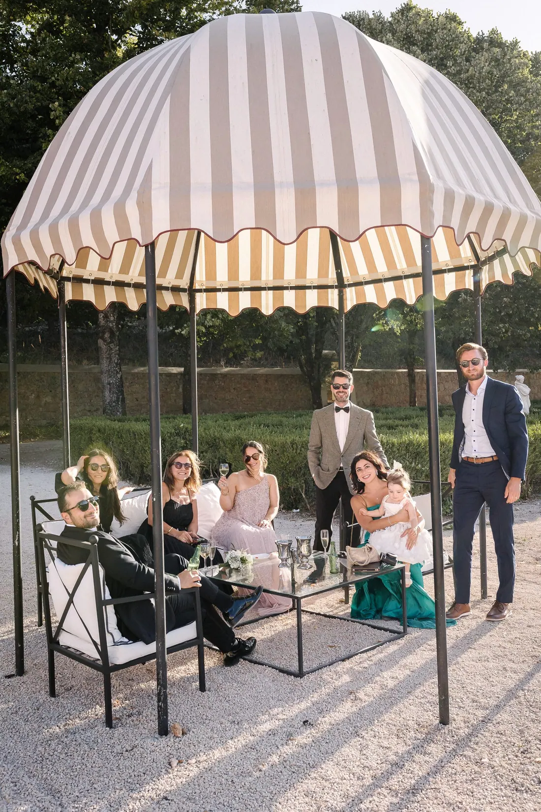 Wedding guests gathered under a striped canopy in a formal garden, sharing a toast