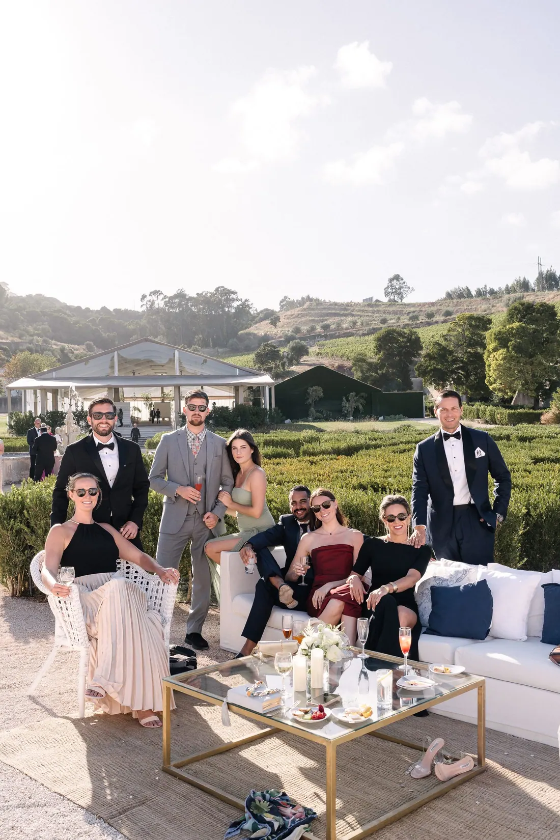 Wedding guests relaxing together in an outdoor lounge with drinks, with vineyards and a glass marquee behind them