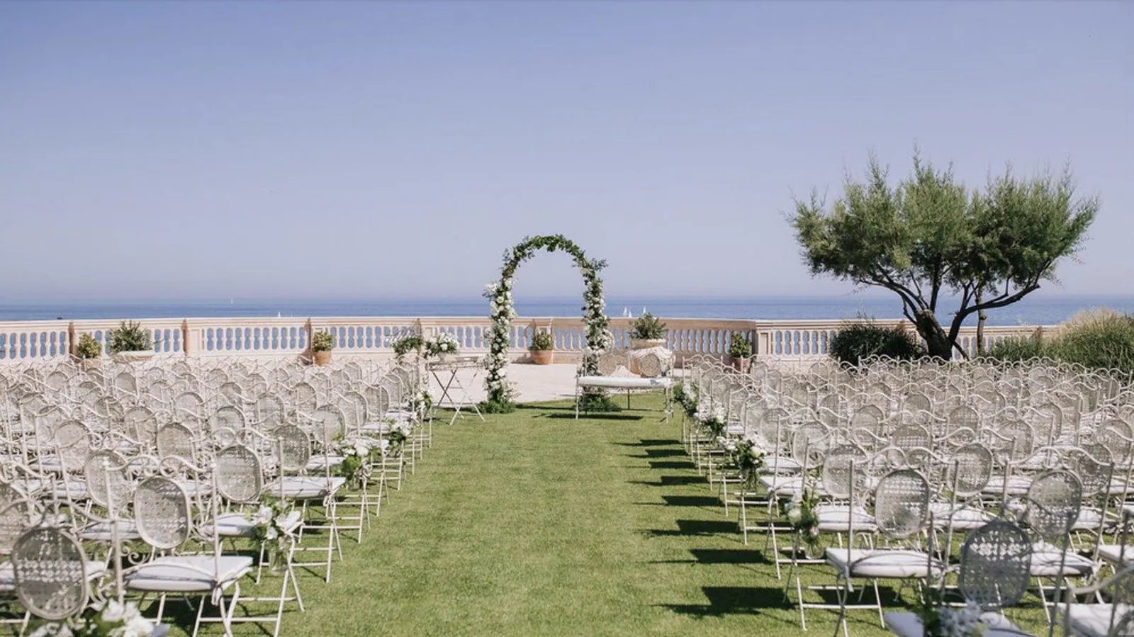 Ceremony set on a seaside terrace lawn, with rows of white chairs facing a floral arch and the sea
